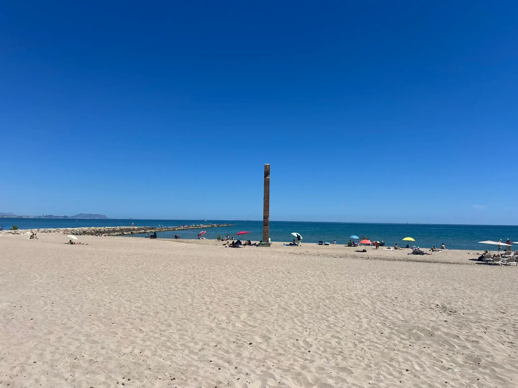 Scenic view of Playa Carrer la Mar in El Campello under a clear blue sky. The wide sandy beach features a tall vertical monument in the center, with scattered beach umbrellas and people enjoying the Mediterranean Sea. A stone breakwater is visible stretching into the calm blue water on the left.